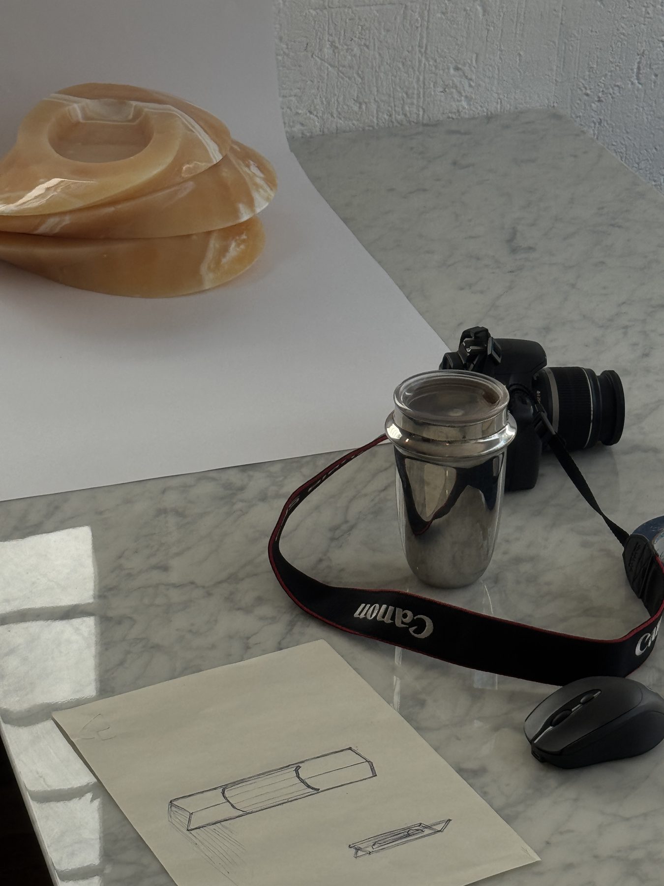 Platos de ónix naranja sobre una mesa de mármol, junto a una cámara, un vaso metálico y un boceto a lápiz